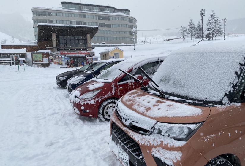 Snow-covered cars parked in front of a modern curved hotel building at a ski resort, with falling snow and ski slopes visible in the background