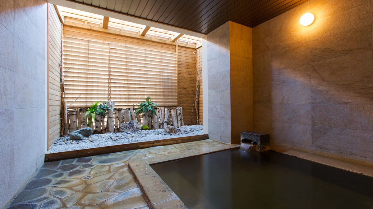 Private onsen bath at Chalet Ivy Niseko featuring dark mineral waters, natural stone flooring, and a Japanese-style window alcove decorated with birch logs, white stones, and green plants under wooden blinds