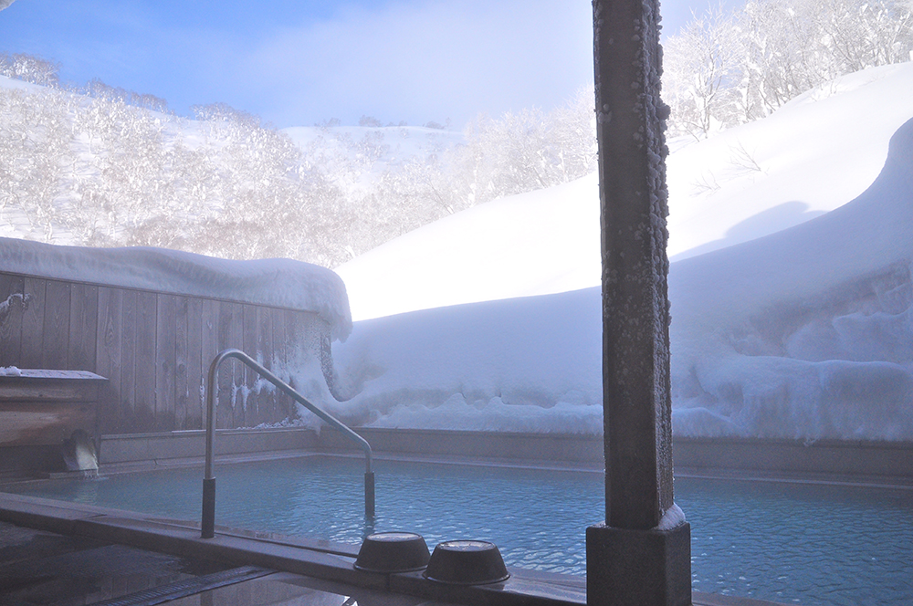 Outdoor onsen bath in winter with steaming blue water surrounded by deep snow banks and frost-covered trees against a bright winter sky, featuring handrails and wooden deck seating