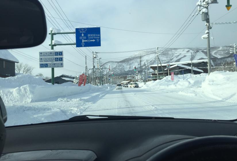 View through a car windshield of a snowy road in Niseko, Japan, with street signs in Japanese and English, power lines, and snow-covered buildings against a mountain backdrop