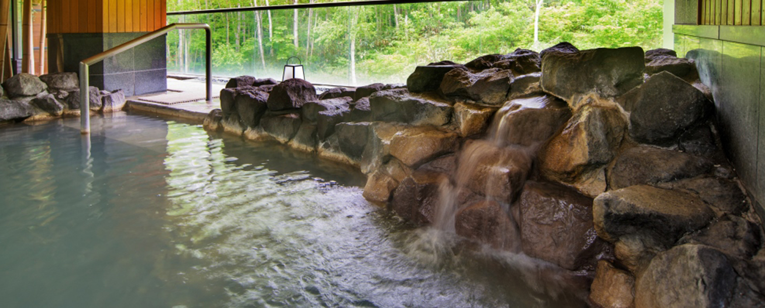 Indoor-outdoor onsen bath at One Niseko Resort Towers featuring cascading water features over natural rock formations, large windows overlooking dense summer forest, and green-tinted mineral waters