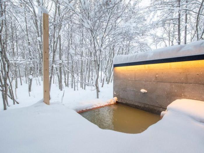 A modern outdoor onsen (Japanese hot spring bath) set in a snowy forest landscape. The bath features a dark overhang with ambient lighting, and is surrounded by snow-covered trees in a serene winter setting