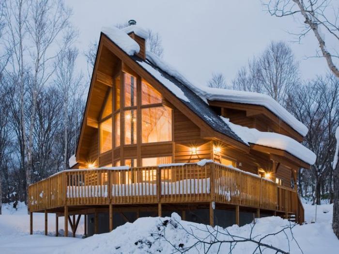 A modern A-frame chalet with large glass windows nestled in a snowy forest setting near Annupuri. The wooden structure features a dramatic peaked roof, expansive deck, and warm interior lighting that glows against the winter twilight backdrop.