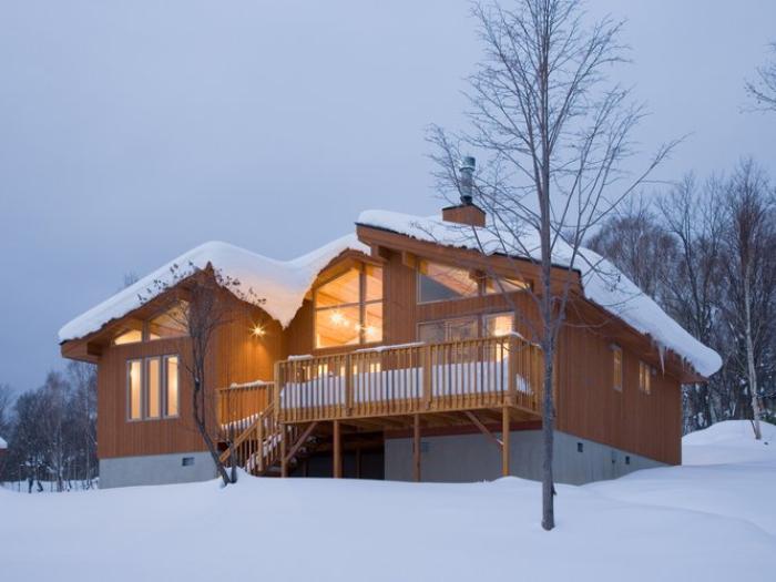 Wood-paneled Chalet Creekside illuminated at dusk, with a wraparound deck, surrounded by snow and bare winter trees in Niseko.