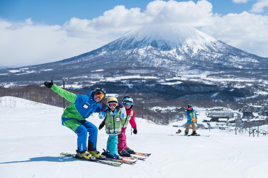 Two children stand with their instructor on a snowy slope with Mount Yotei in the back ground