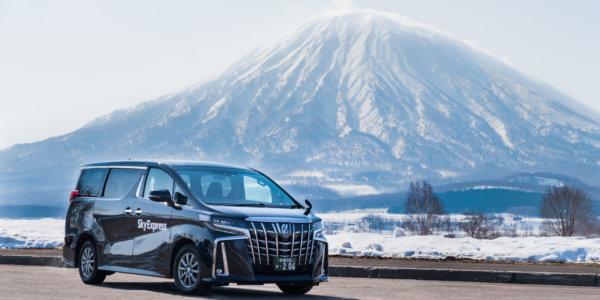 A black Alphard van in a car park with a snow covered Mount Yotei behind.