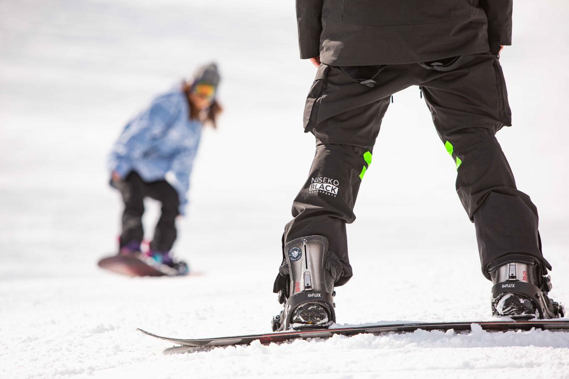 A view of a snowboarder in front of the legs of their instructor