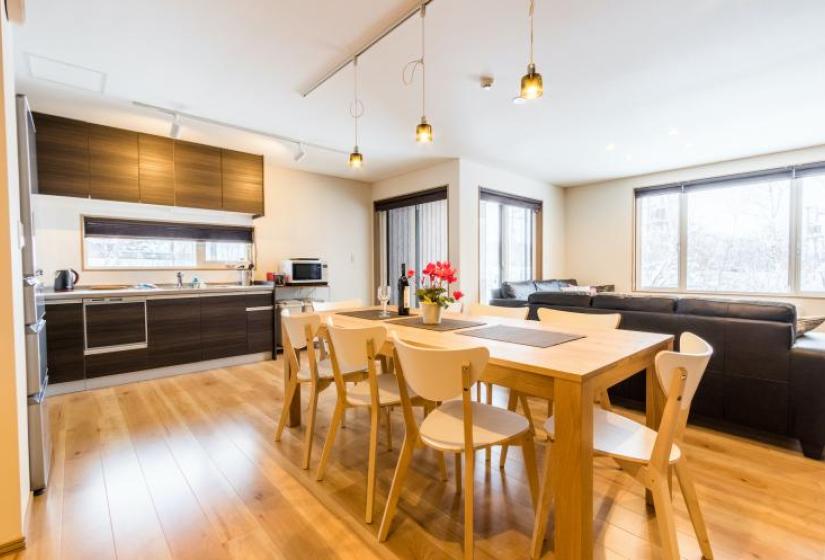 A dining table and kitchen behind with hanging pendant lights