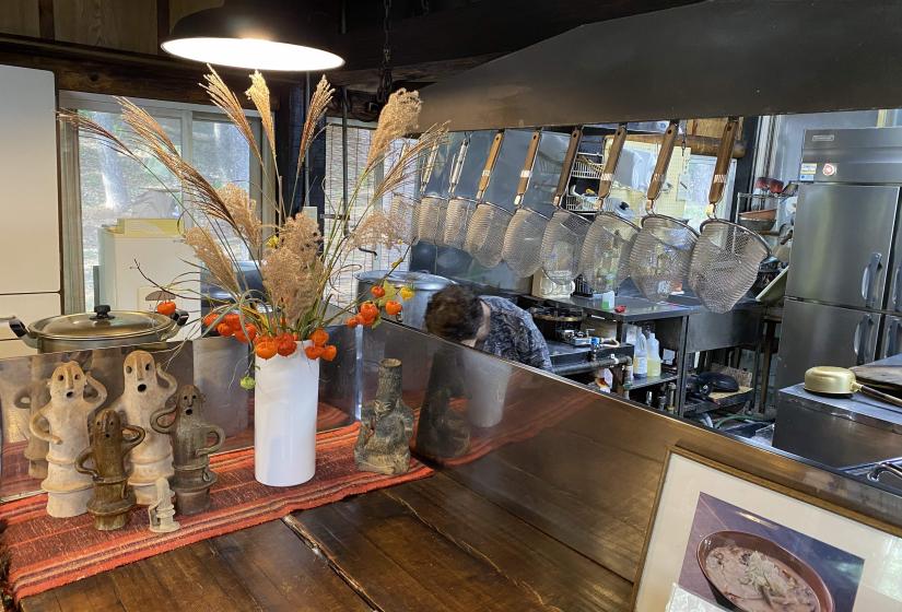 A view into a ramen shop kitchen with hanging noodle sieves and a dried flower arrangement on a bench.