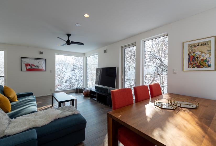 Dining table with red chairs and television and large windows behind.
