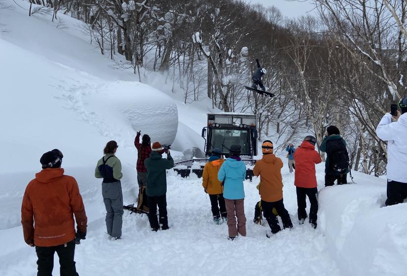 A snowboarder jumps over a snow cat in front of onlookers