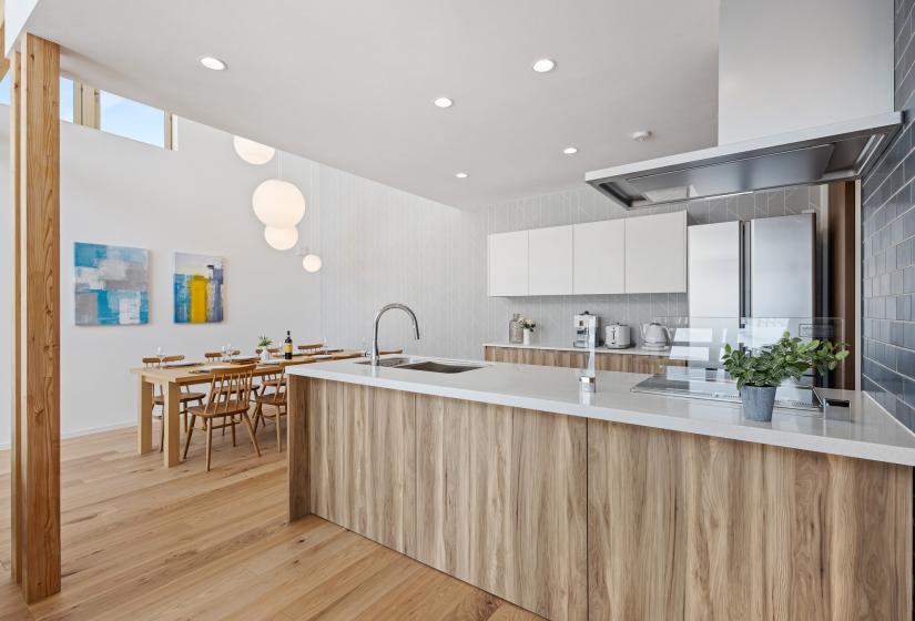 Modern open-plan kitchen and dining space with light wood cabinetry, white countertops, and stainless steel appliances. Natural light streams in through high windows, illuminating the wooden dining table and chairs in the background