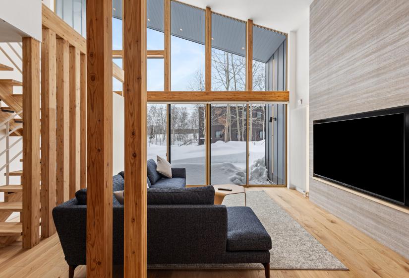 Contemporary living room with floor-to-ceiling windows showcasing Niseko's winter landscape. Features modern black sofas, wooden architectural details, and a mounted flatscreen TV against a feature wall