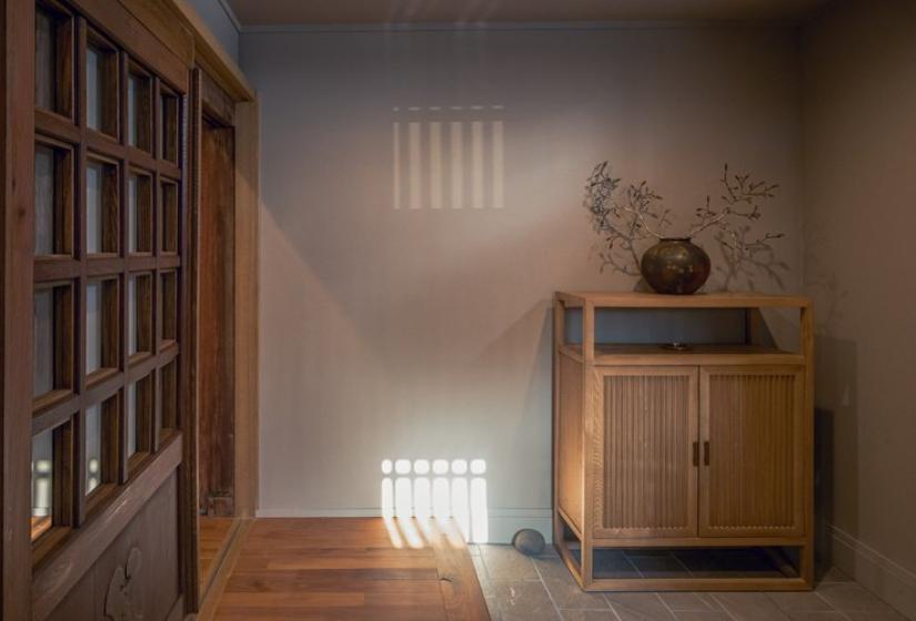Serene corner at Nagare House showcasing traditional Japanese design with wooden latticed windows casting geometric shadows on the neutral wall, a wooden cabinet with minimalist decor, and polished wooden flooring. This authentic detail from the restored 100-year-old kominka farmhouse captures the traditional Japanese aesthetic that makes this luxury Niseko ski accommodation unique.