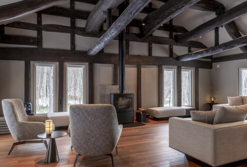 Inviting lounge area at Nagare House featuring comfortable beige armchairs arranged around a central wood-burning fireplace, exposed traditional wooden beam ceiling structure, polished wooden floors, and large windows bringing in natural light. This warm gathering space in the restored 100-year-old kominka farmhouse offers the perfect setting for après-ski relaxation in luxury Niseko accommodation.