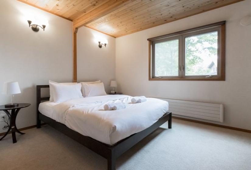 A clean, minimalist bedroom with a queen bed featuring white linens and dark wooden frame. The room has light-colored carpeting, white walls, wooden ceiling, a small window with natural light, bedside table, and wall sconces creating a simple and peaceful sleeping space.