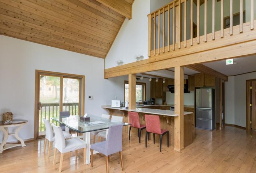 An open-concept dining and kitchen area with wooden floors and ceiling. The space features a white dining table with chairs, a kitchen island with bar stools, and a loft area above. Large windows provide natural light, and the layout showcases the chalet's modern yet warm interior design.