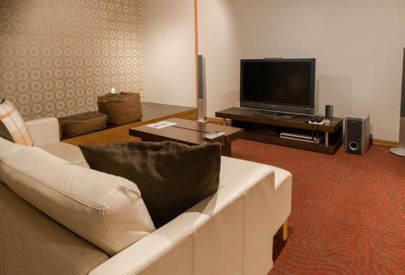 A cozy indoor living area with a white sofa, wooden coffee table, and entertainment center featuring a flat-screen TV. The room has a warm atmosphere with red carpeting, wooden furnishings, and neutral walls creating a comfortable space for relaxation.