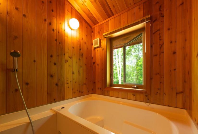 A traditional Japanese-style bath with wooden paneled walls and ceiling. The bathroom features a deep soaking tub, window with forest views, and warm lighting creating a serene bathing space with natural elements.