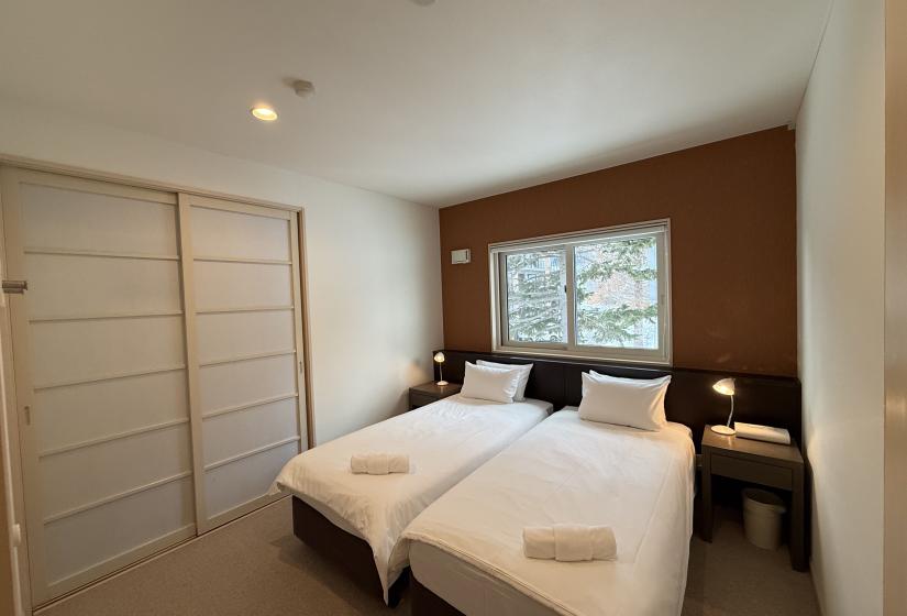Bright twin bedroom at Yutaka Townhouses featuring two single beds with fresh white linens, wood-accented walls, large windows with forest views, and built-in closet space for winter ski holiday stays.
