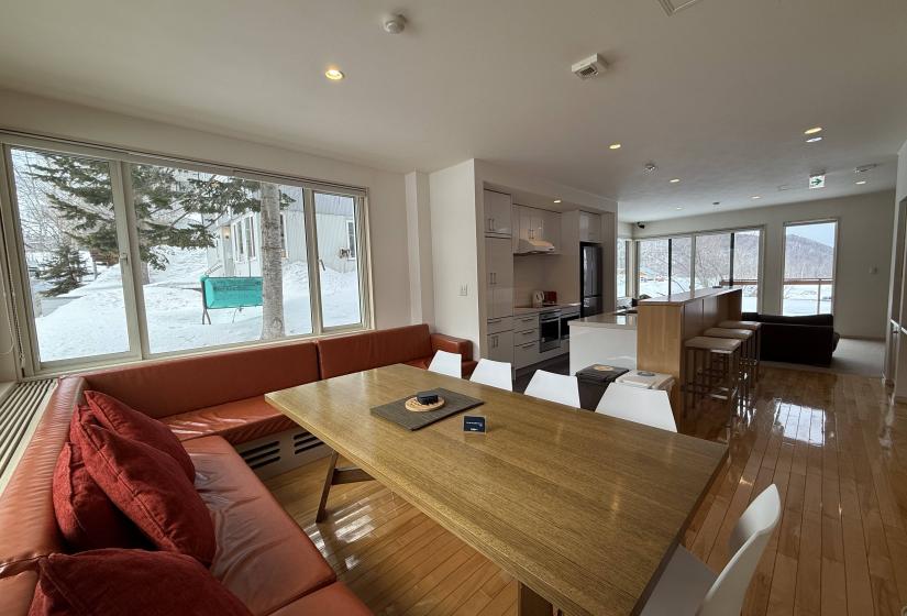 Bright dining area at Yutaka Townhouses featuring solid wooden dining table with red bench seating, large windows overlooking snowy Niseko landscape, and open connection to living room and kitchen, ideal for family meals during ski holidays.