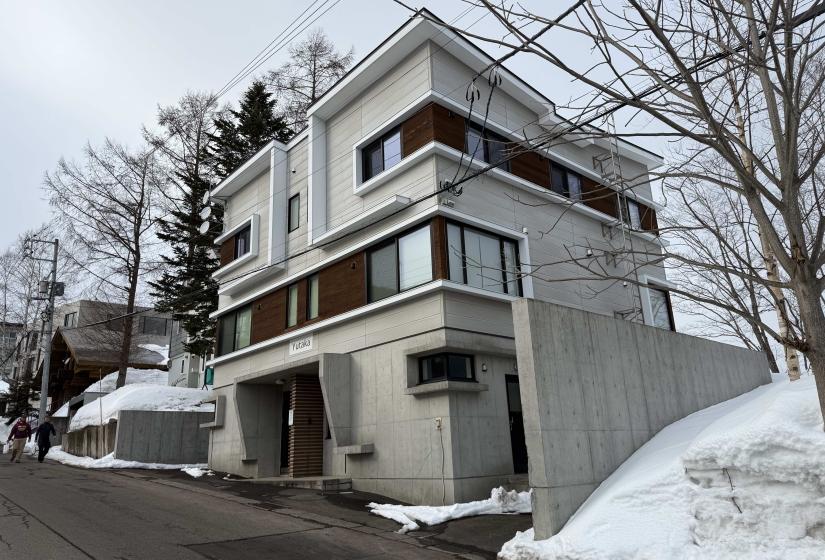 Exterior view of modern Yutaka Townhouses in Niseko, featuring contemporary architecture with wooden accents, surrounded by snow-covered landscape, within walking distance to Hirafu ski lifts.