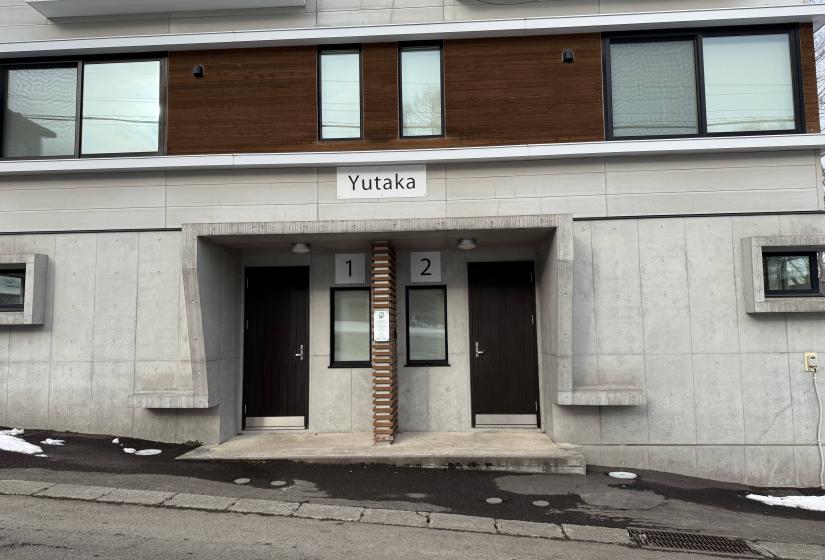 Front entrance of Yutaka Townhouses showing modern concrete and wood facade, twin entry doors, property name sign, and snowy street access, centrally located in Niseko for convenient ski holiday accommodation.