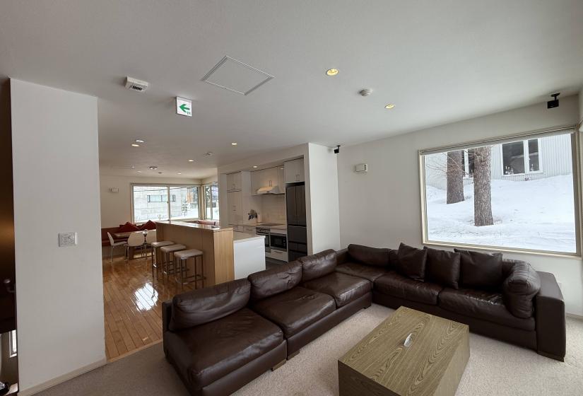 Open-concept living space at Yutaka Townhouses featuring plush dark leather sectional sofa, coffee table, large windows with snow views, and seamless flow to dining and kitchen areas, ideal for group gatherings during Niseko ski holidays.