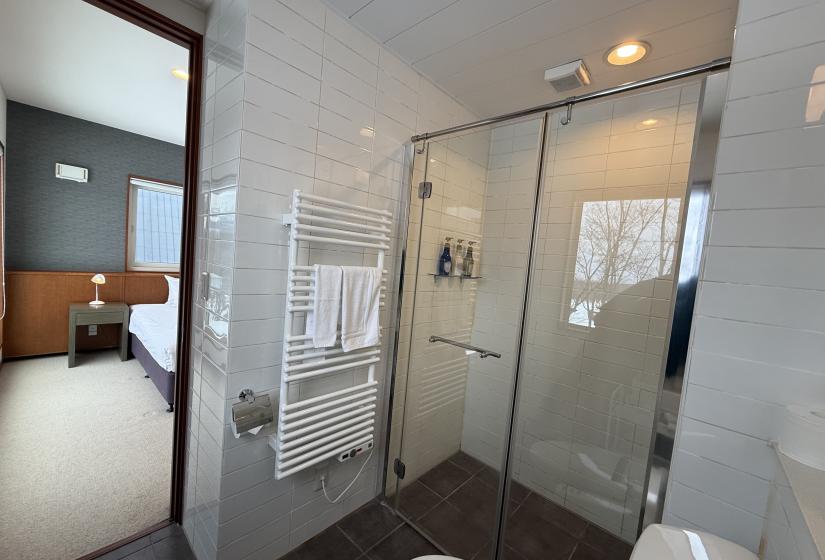 Modern bathroom at Yutaka Townhouses featuring glass shower enclosure, heated towel rack, white tiled walls, and doorway leading to bedroom area, providing premium amenities for Niseko ski holiday guests.