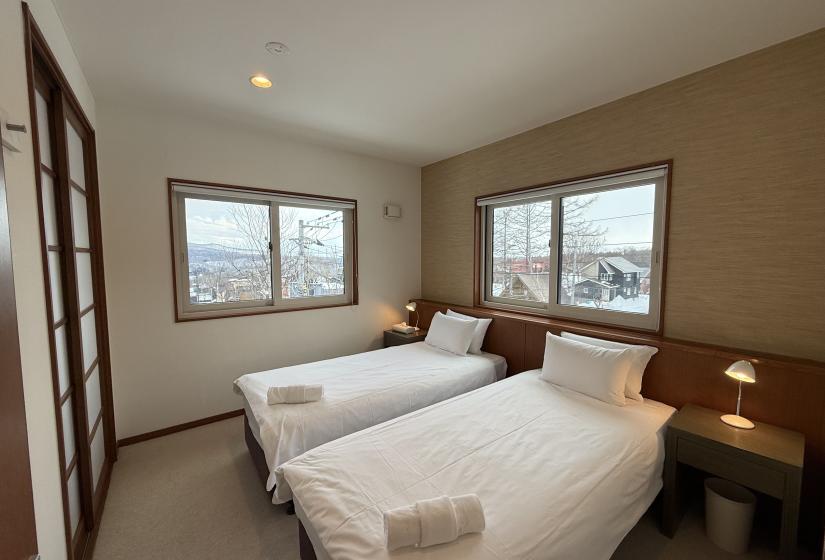 Twin bedroom at Yutaka Townhouses with two comfortable beds, warm wood accent walls, dual windows offering mountain and village views, bedside tables with reading lamps, and neutral decor creating a relaxing retreat after skiing in Niseko.