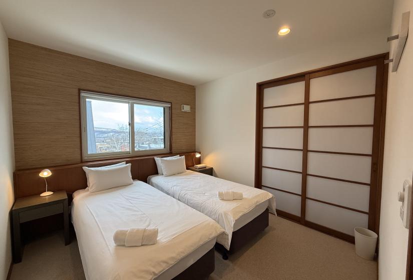 Twin bedroom at Yutaka Townhouses featuring two plush beds with crisp white linens, Japanese-style sliding closet doors, wooden accent wall, large window with mountain views, and bedside lamps for cozy Niseko ski holiday accommodation.