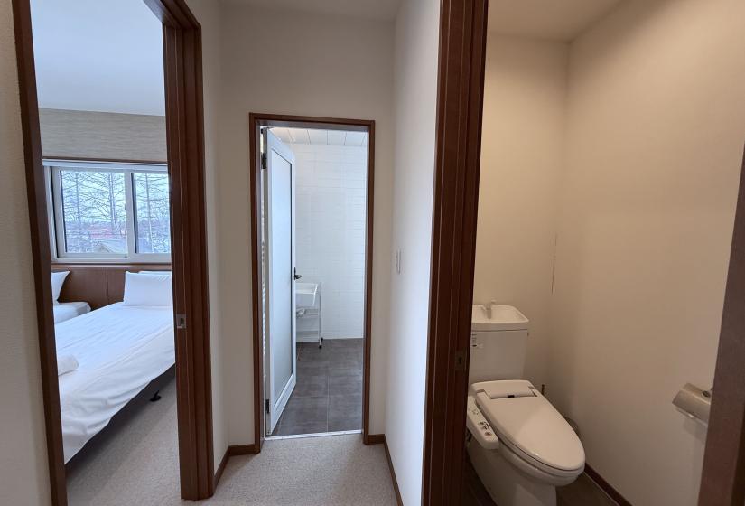 Hallway at Yutaka Townhouses showing doorway to bedroom with white bedding, entrance to modern bathroom with toilet visible, and clean contemporary design with wooden door frames, providing convenient layout for guests during their Niseko ski holiday.
