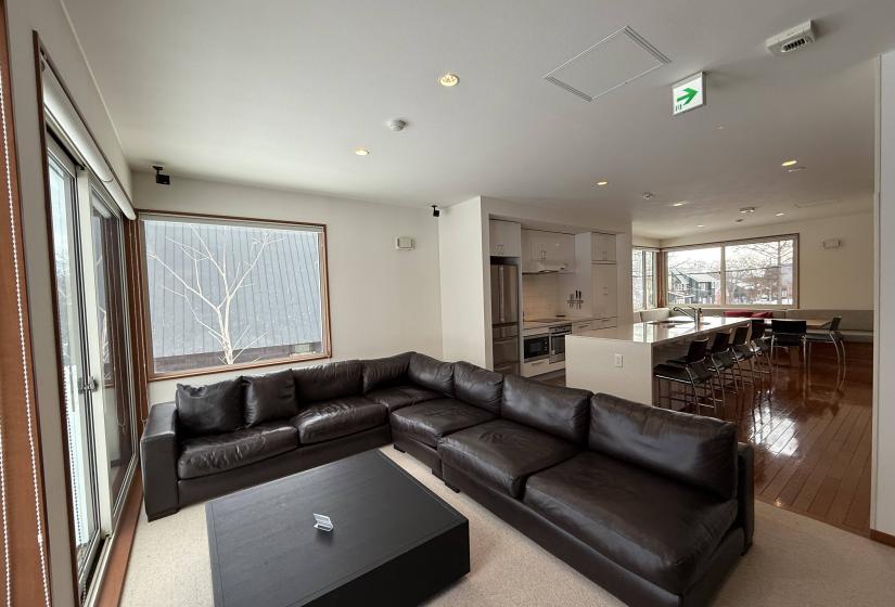 Spacious living room at Yutaka Townhouses featuring large black leather sectional sofa, coffee table, bright windows with forest views, and open-plan layout connecting to dining area, offering comfortable relaxation space for groups enjoying Niseko ski holidays.