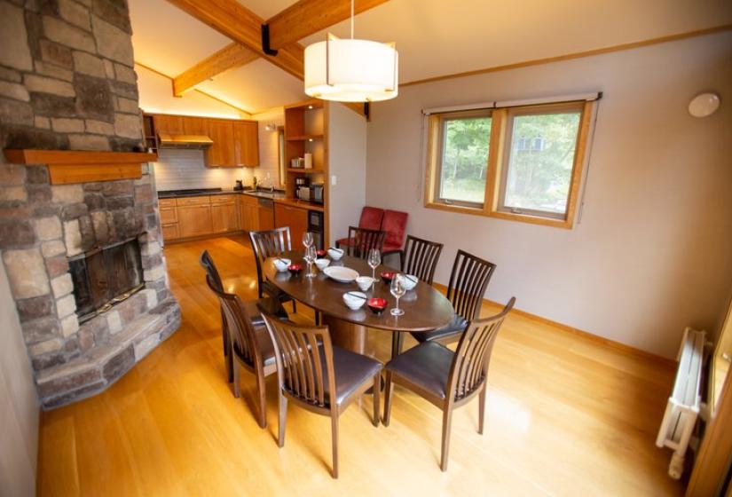Dining area with glass table and leather chairs, wooden flooring, stone accent wall, pendant lighting, and windows with forest views at Chalet Creekside.