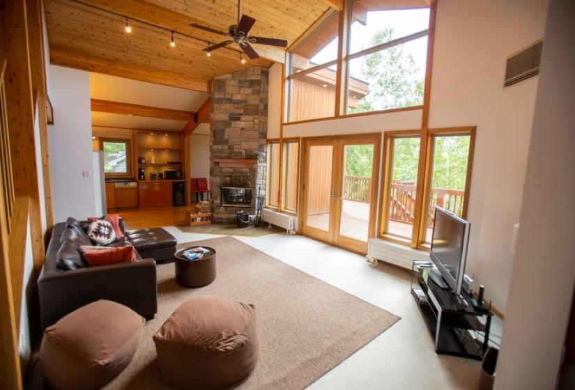 Large living room with wooden ceiling, stone fireplace, comfortable couches, floor-to-ceiling windows, and natural light at Chalet Creekside.