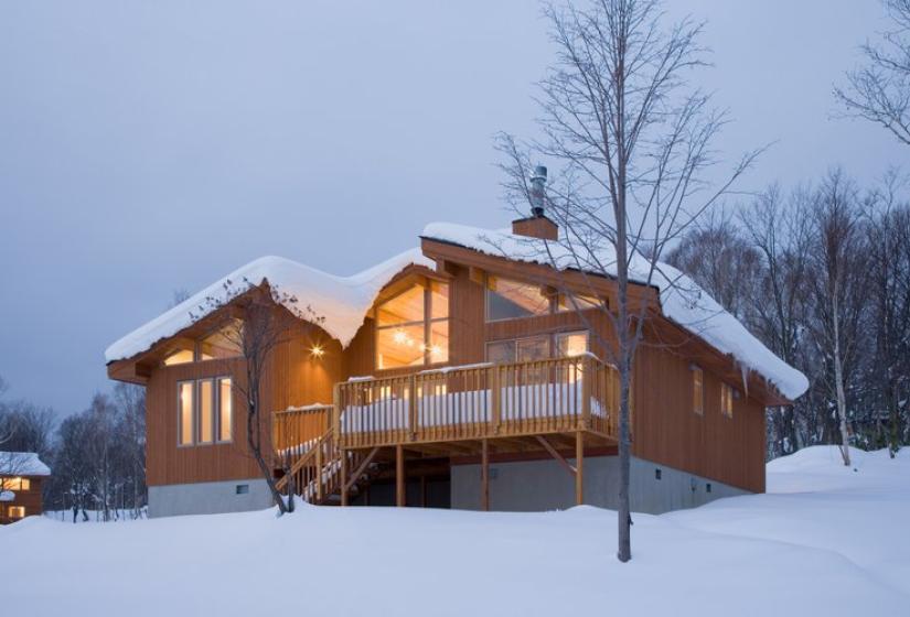 Wood-paneled Chalet Creekside illuminated at dusk, with a wraparound deck, surrounded by snow and bare winter trees in Niseko.