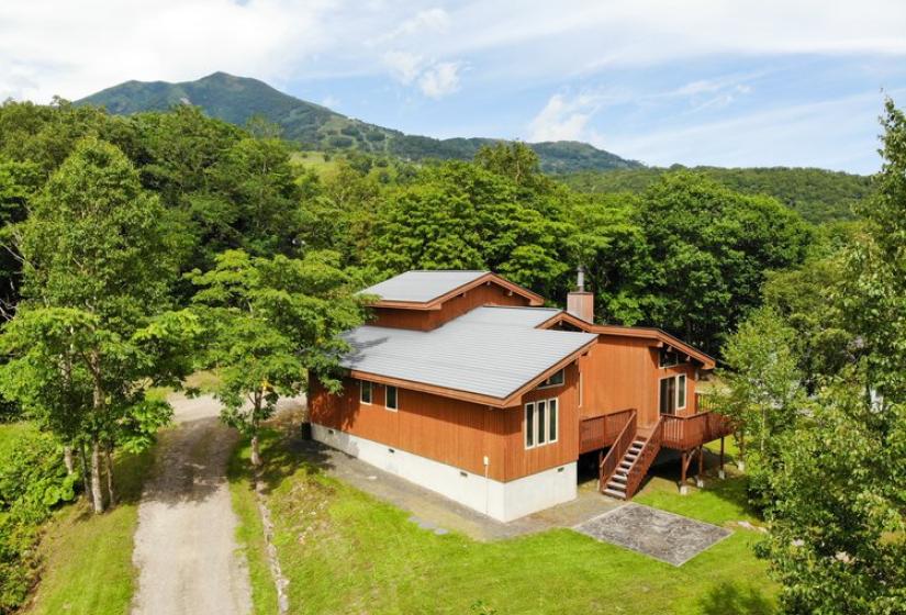 Aerial view of Chalet Creekside surrounded by lush green forest and mountains in summer, showing the wooden structure with metal roof and surrounding landscape.