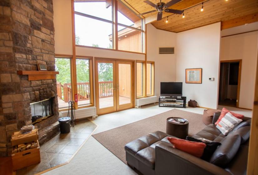 Large living room with high ceiling, stone fireplace, gray sectional sofa, wooden beams, and floor-to-ceiling windows overlooking greenery at Chalet Creekside.