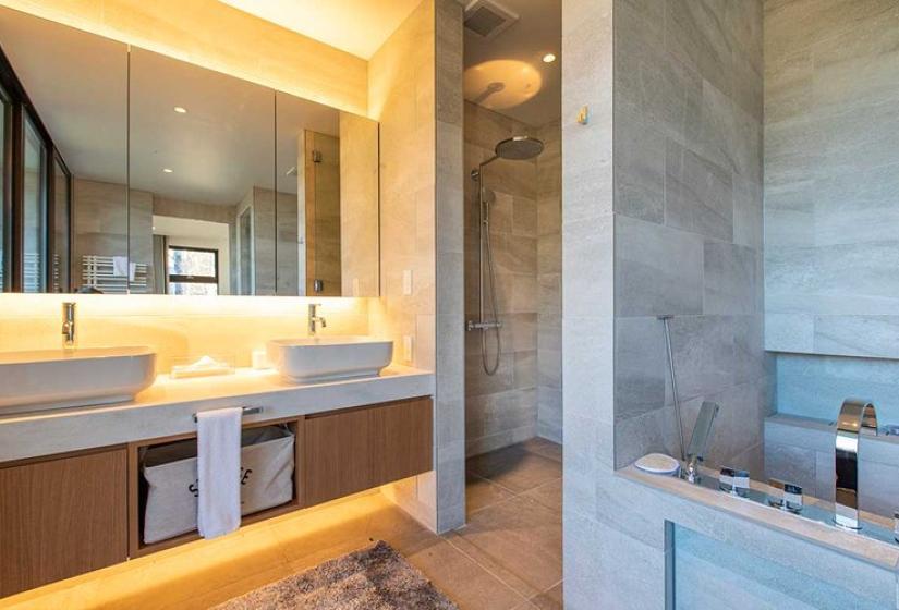 Elegant master bathroom in luxury Niseko ski chalet featuring dual vessel sinks with wooden vanity, ambient LED lighting, stone tile walls, glass shower enclosure, and deep soaking tub with forest views - offering premium après-ski relaxation in Japan's premier winter destination.