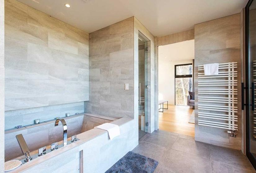 Luxurious master bathroom in premium Niseko ski chalet featuring deep soaking tub, modern stone tile walls, glass shower enclosure, heated towel rack, and natural light from large windows - offering spa-quality relaxation after days skiing Japan's world-famous powder.