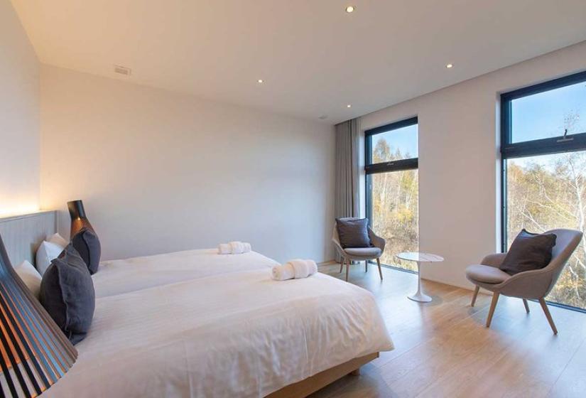 Bright twin bedroom in luxury Niseko ski chalet featuring two comfortable beds with crisp white linens, dark blue accent pillows, light wood flooring, modern accent chairs, and large windows framing serene forest views - ideal for restful recovery after skiing Japan's world-famous powder.