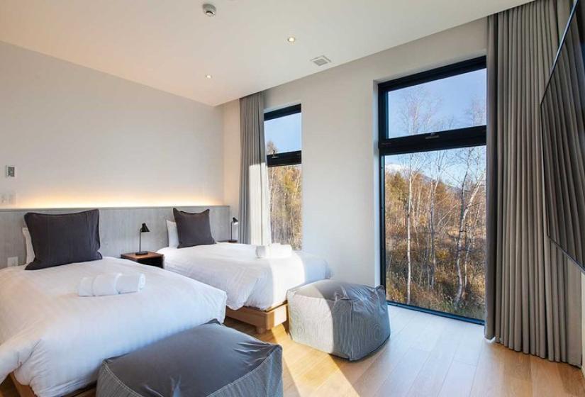 Elegant bedroom in luxury Niseko ski chalet featuring two comfortable beds with premium white linens, soft gray headboards, modern pendant lighting, light wood flooring, and floor-to-ceiling windows showcasing stunning birch forest views - perfect for restful nights after skiing Japan's famous powder.