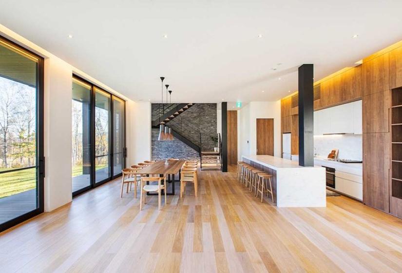 Spacious open-concept living area in luxury Niseko ski chalet featuring light wood flooring, large wooden dining table, contemporary kitchen with white island, sleek black structural columns, floor-to-ceiling windows overlooking nature, and warm ambient lighting - offering a perfect blend of Japanese minimalism and alpine luxury.
