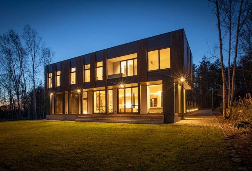 Stunning twilight exterior view of a luxury modern ski chalet in Niseko featuring dark wood cladding, dramatic floor-to-ceiling windows illuminated from within, contemporary architectural design, expansive lawn area, and surrounded by birch trees - showcasing premium Japanese alpine accommodation at its finest.