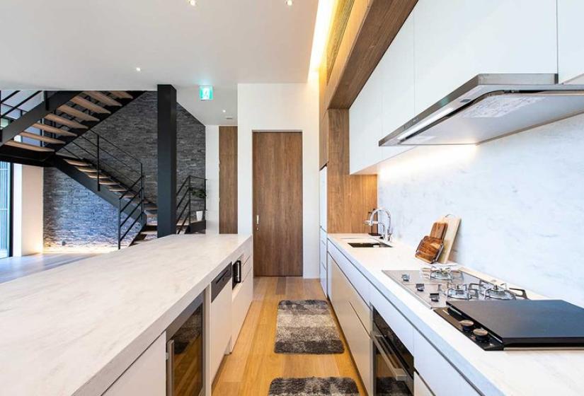 Designer kitchen in luxury Niseko ski chalet featuring sleek white countertops, high-end integrated appliances, professional gas cooktop, minimalist cabinetry, warm wood accents, and open layout with view to modern staircase - perfect for culinary experiences during premium Japanese ski holidays.