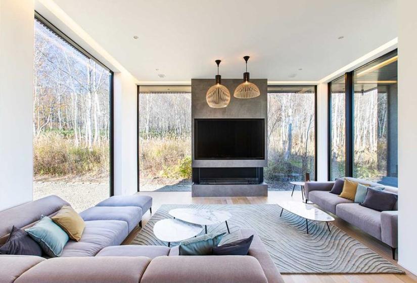 Modern living room in luxury Niseko ski chalet featuring comfortable light gray sectional sofas, contemporary glass coffee tables, designer pendant lighting, large flat-screen TV with stone feature wall, patterned area rug, and floor-to-ceiling windows offering panoramic forest views - an ideal space for relaxation after experiencing Japan's legendary powder snow.