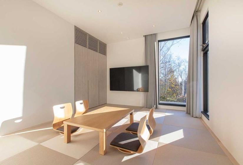 Minimalist Japanese-inspired tatami room in luxury Niseko ski chalet featuring traditional low wooden table, wall-mounted TV, large window with forest views, light wood flooring, and simple elegant design - offering authentic cultural experience within an upscale ski accommodation in Japan.