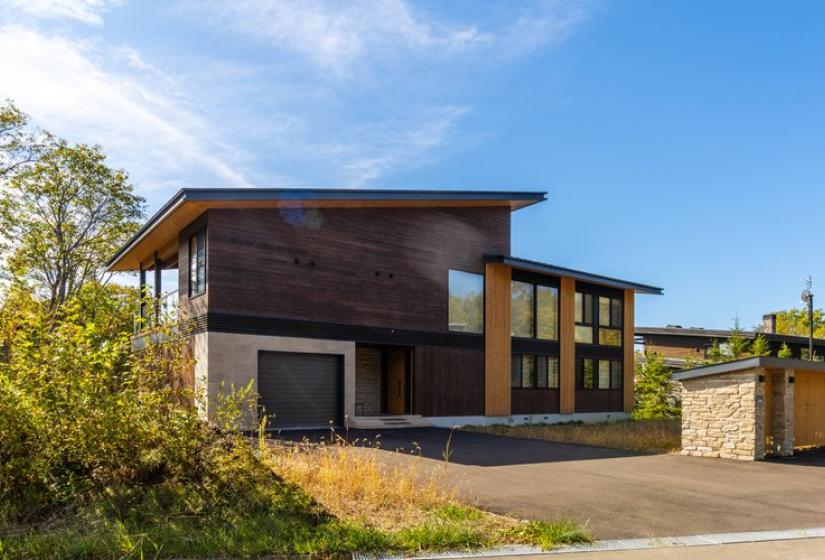 Exterior view of Chalet Lotus Niseko showcasing its contemporary architectural design with dark wood cladding, angled roof lines, large windows, and natural stone elements, perfectly positioned in Niseko's stunning alpine landscape for luxury ski accommodation.