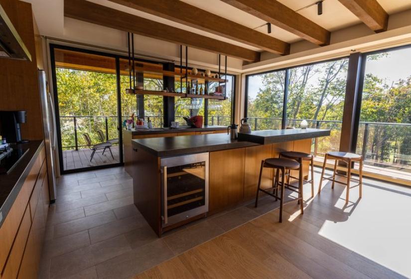 Modern open-concept kitchen in Chalet Lotus Niseko featuring a central island with wooden breakfast bar, exposed ceiling beams, floor-to-ceiling windows overlooking lush forest, and contemporary design perfect for luxury alpine getaways.