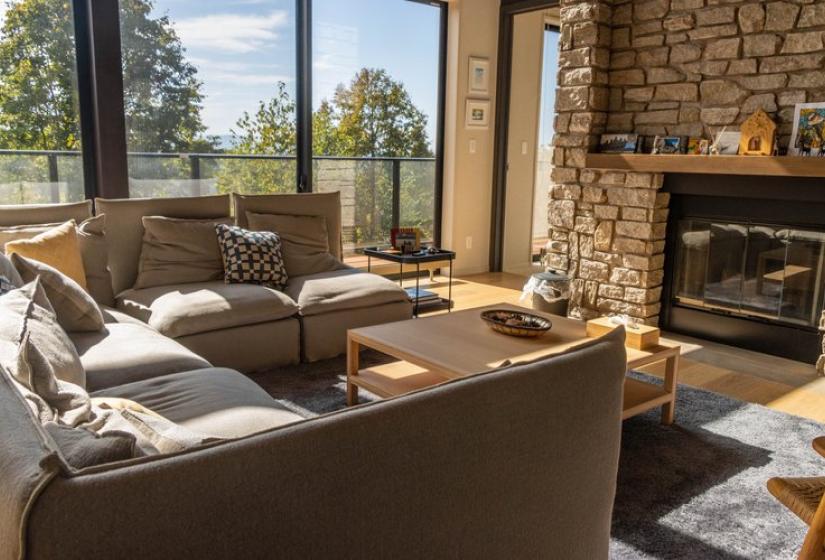Inviting living room at Chalet Lotus Niseko featuring comfortable sectional sofa, stone fireplace, wooden coffee table, floor-to-ceiling windows with mountain scenery, and natural light creating a perfect luxury alpine retreat atmosphere.